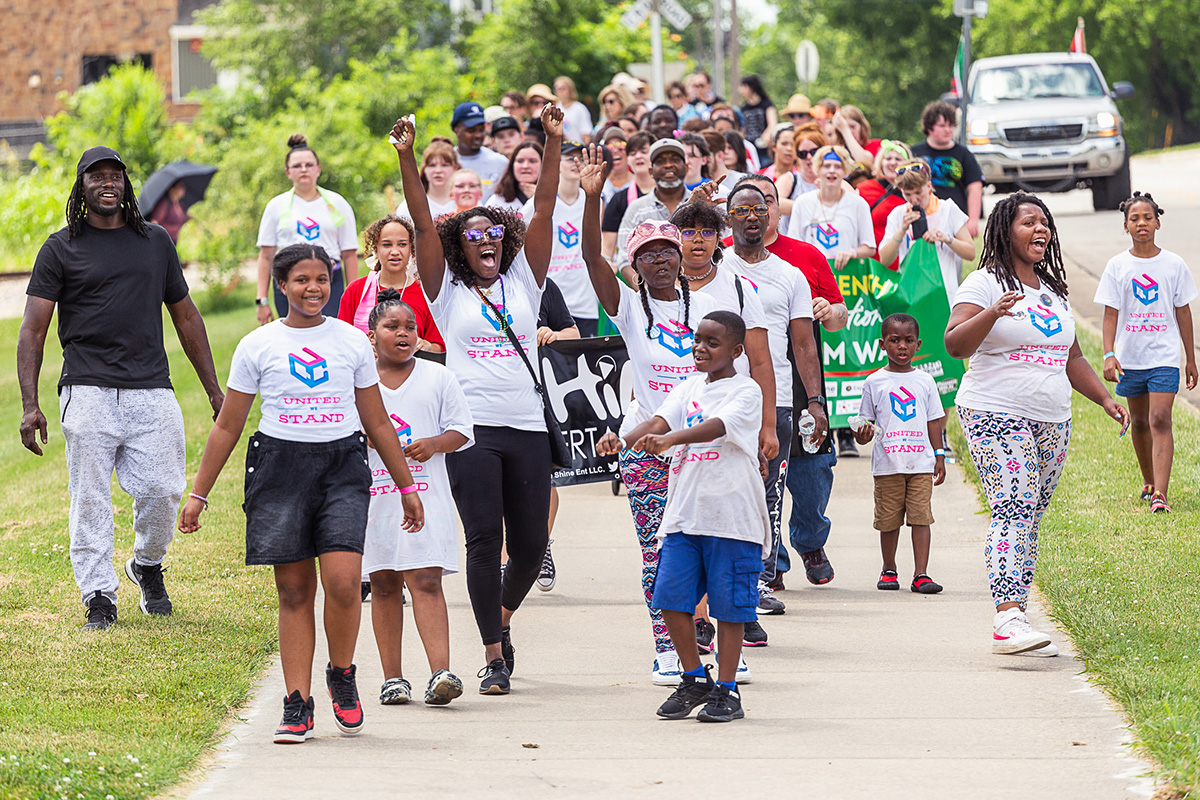 A group of people marches joyfully down a sidewalk. Many wear white t-shirts that say UNITED WE STAND.