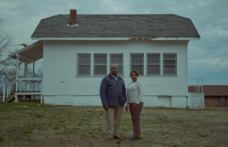 Two people pose in front of a weathered-looking schoolhouse.