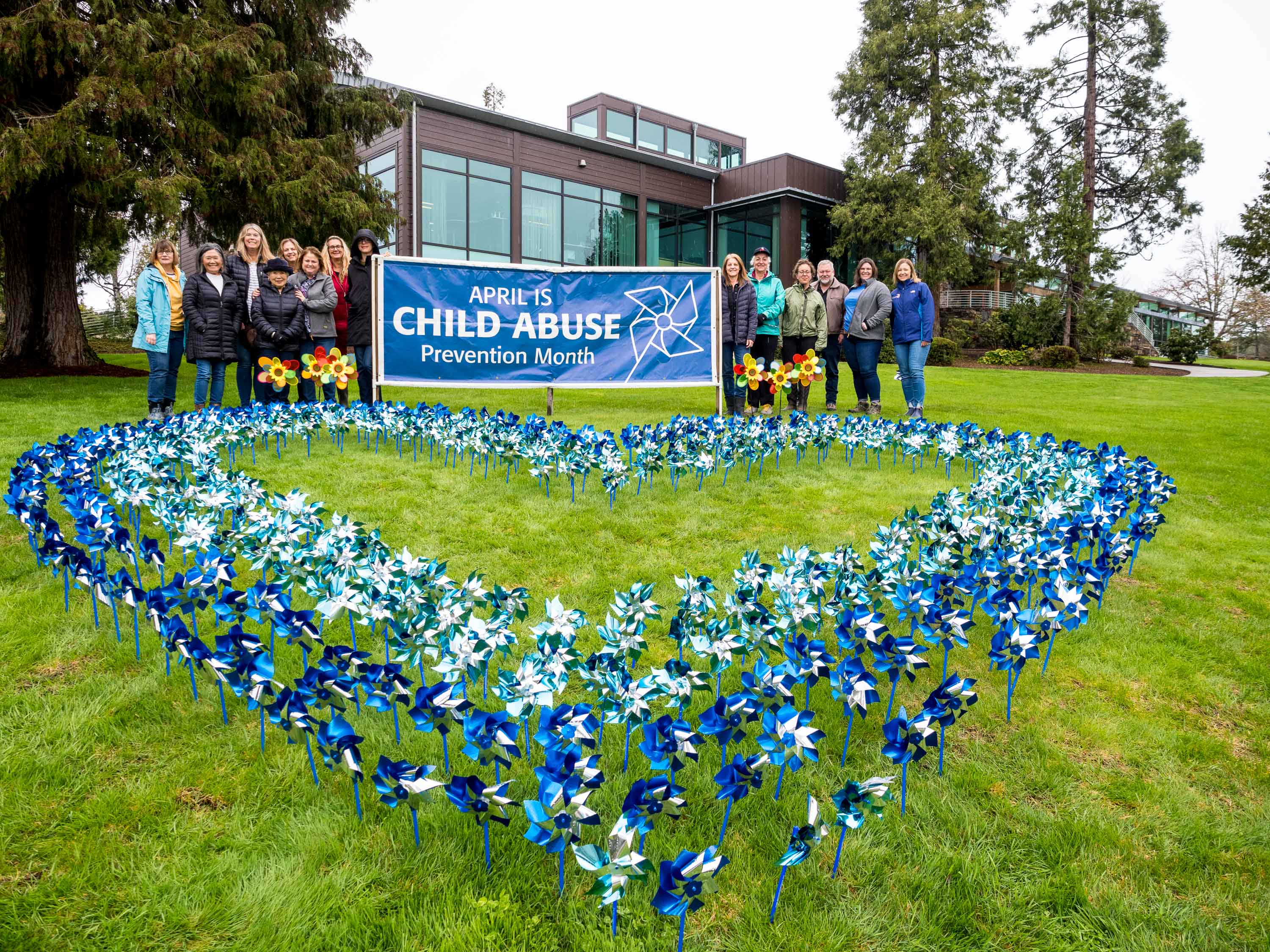 A group of people stands behind a large heart made of pinwheels planted into grass. A banner reads April is Child Abuse Prevention Month.