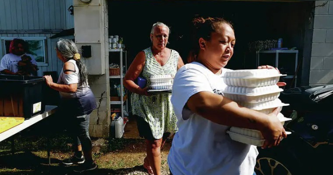 JAMM AQUINO / JAQUINO@STARADVERTISER.COM  Kim Fanene, right, co-founder of Farm Kine Tings, and Kahana Bay resident Debra Gorai, middle, carry trays of hot food for distribution, Thursday, Aug. 20, 2026, in Punaluu.