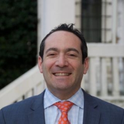 Jonathan Katz wears a blue shirt, navy jacket, and red tie and stands in front of a white building