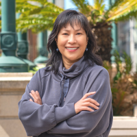 May Leong, an Asian American woman wearing a gray jacket against a brightly colored background