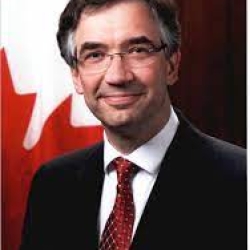 Roman Waschuk wears a dark suit with a red tie and stands in front of the Canadian flag